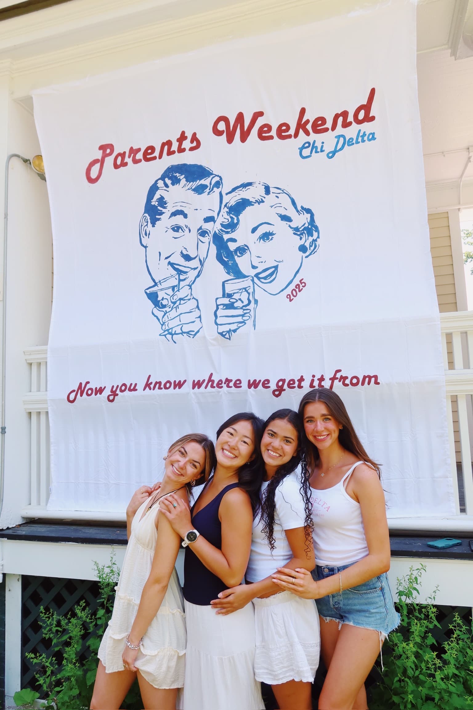 Four sisters in front of the hand-painted Parents Weekend banner on the haus porch