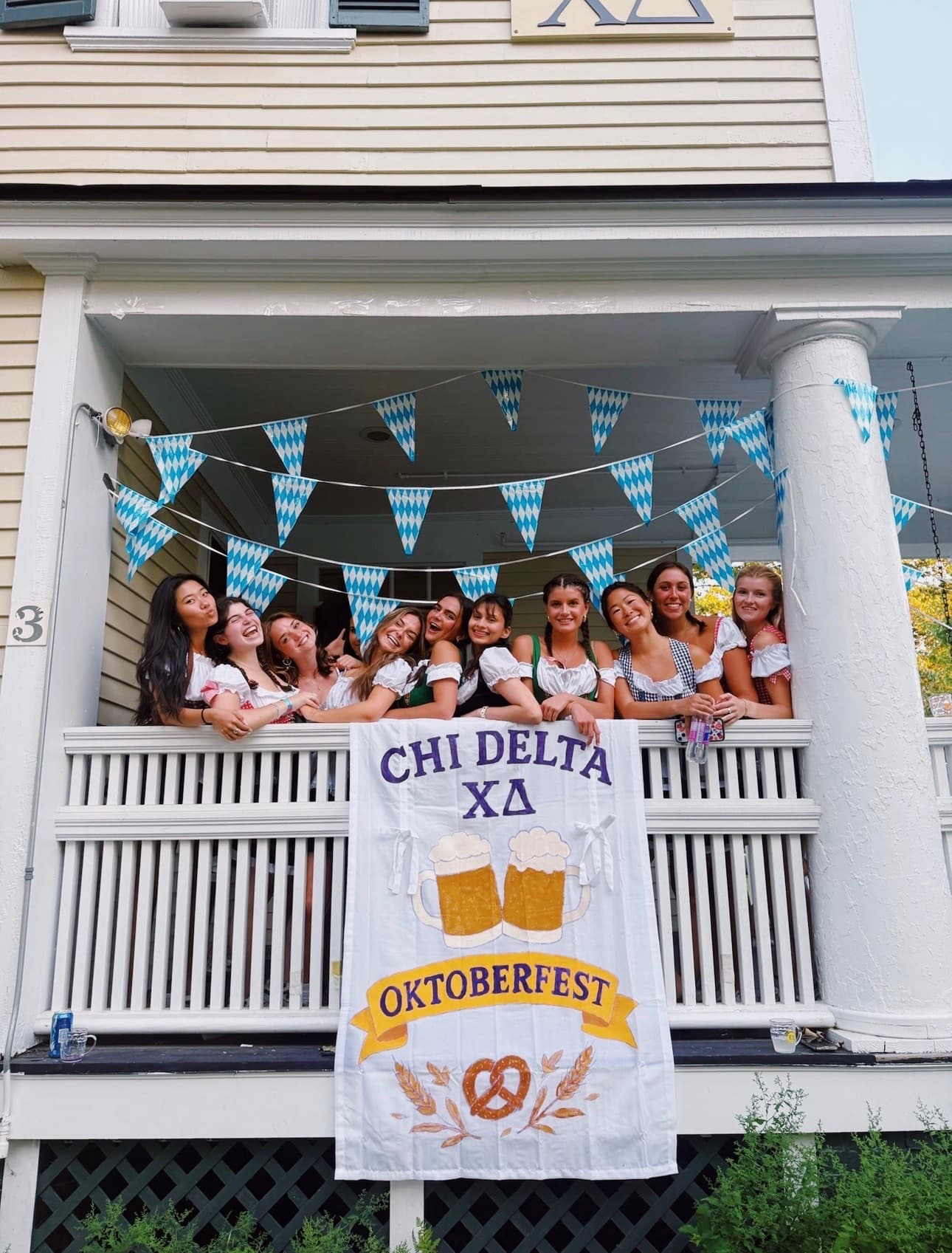 Sisters in dirndls leaning over the haus porch railing above the hand-painted Oktoberfest banner