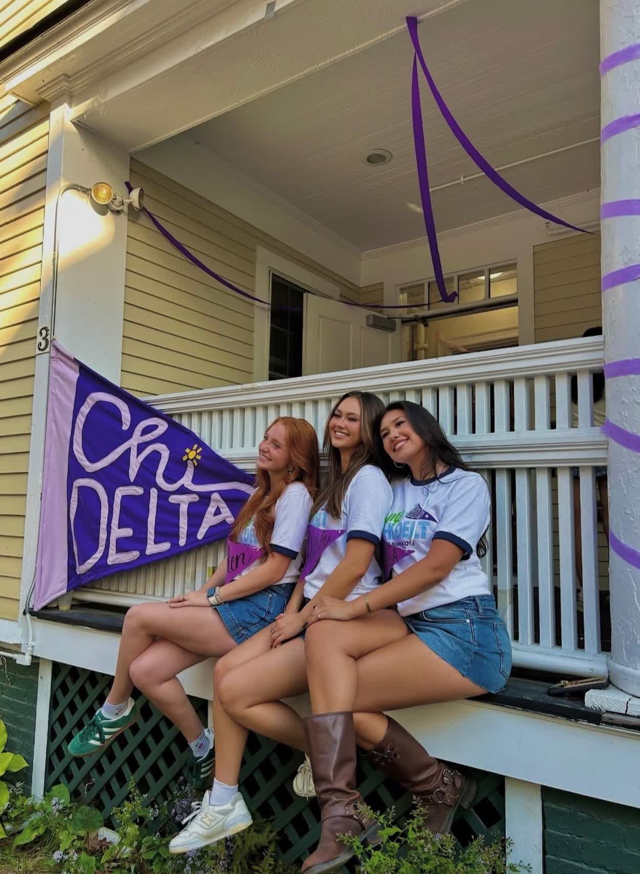 Three sisters on the porch railing beside the hand-painted Chi Delta pennant